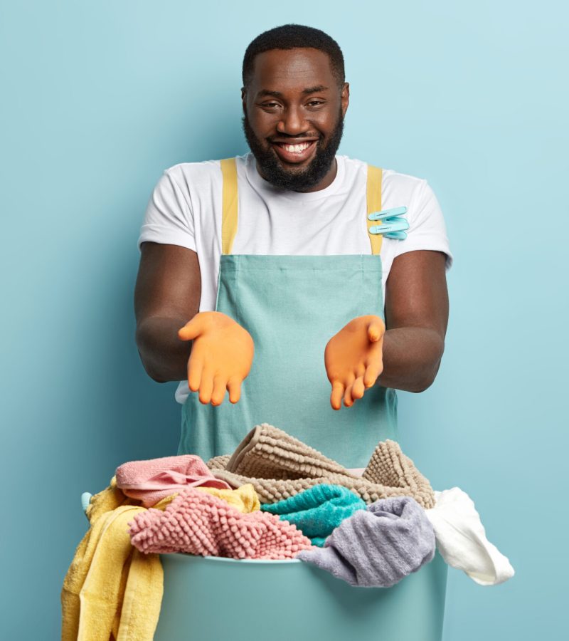 Cheerful householder with dark skin demonstrates washed clean garment, keeps hands near basin with tidy laundry, satisfied with quick good result of work, going to fasten wet clothes on line